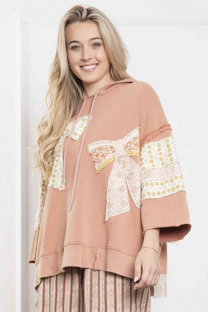 A smiling woman wears a terracotta-colored hoodie with 3/4 sleeves and contrast patchwork detailing in floral and lace fabrics. The hoodie features a drawstring hood and oversized fit with raw hem accents, styled with wide-leg striped pants in coordinating tones.