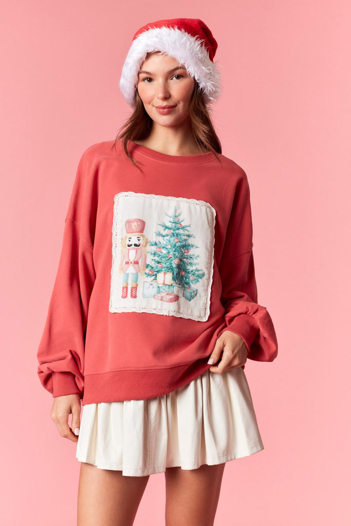 Dark coral Christmas sweatshirt featuring a nutcracker and Christmas tree corduroy patch with scalloped lace trim, styled with a pleated skirt and Santa hat.