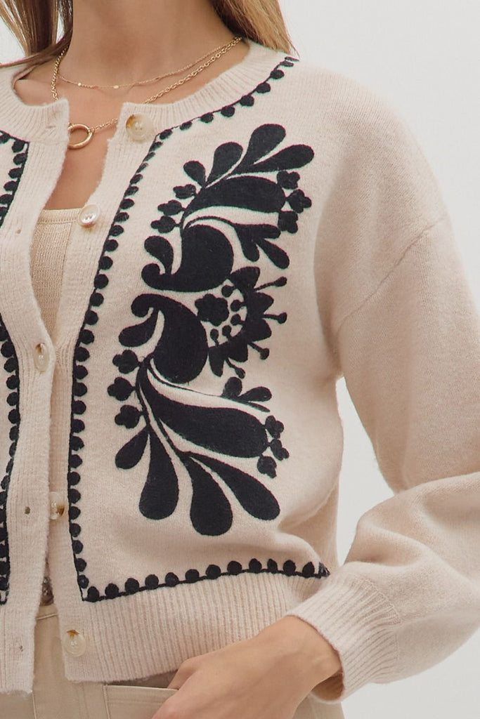 An oatmeal-colored sweater knit cardigan with black floral embroidery on the front panels and a scalloped trim on the crew neckline, hem, and button placket.