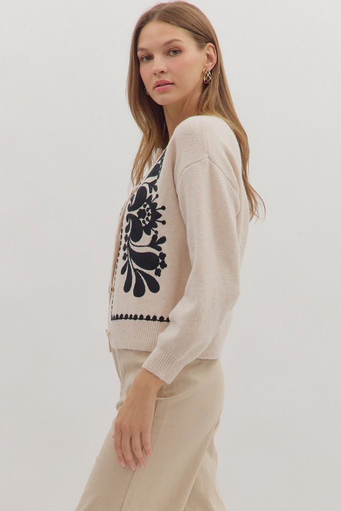 An oatmeal-colored sweater knit cardigan with black floral embroidery on the front panels and a scalloped trim on the crew neckline, hem, and button placket.