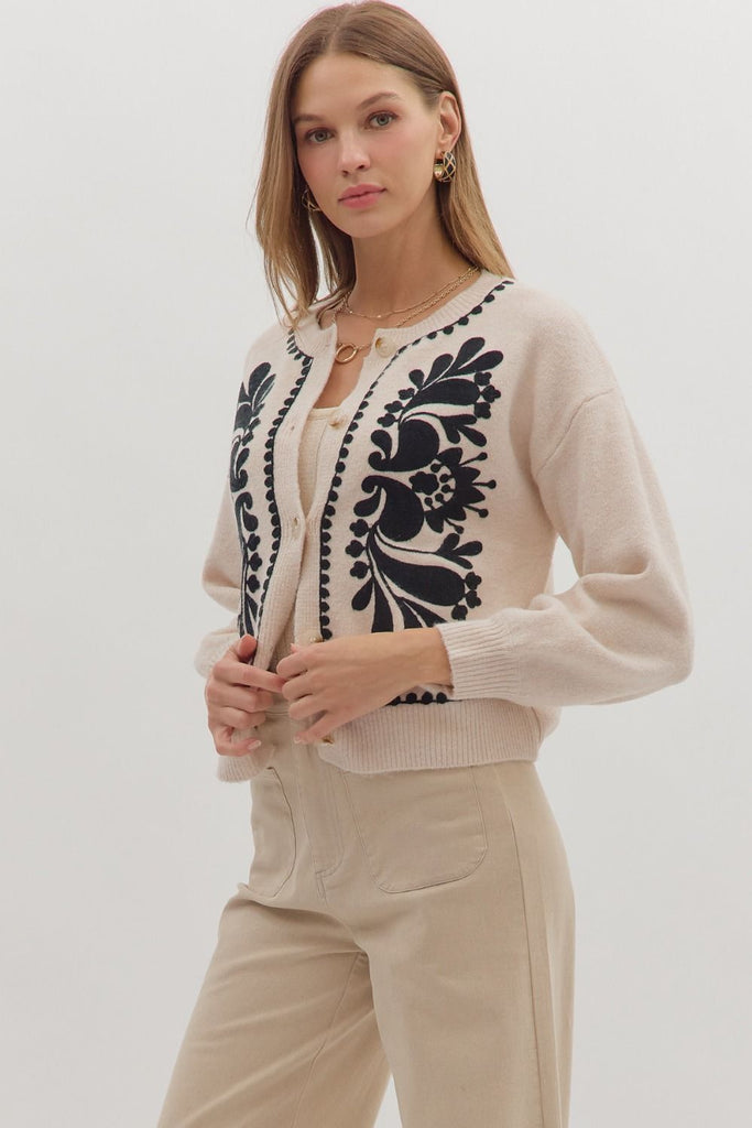 An oatmeal-colored sweater knit cardigan with black floral embroidery on the front panels and a scalloped trim on the crew neckline, hem, and button placket.