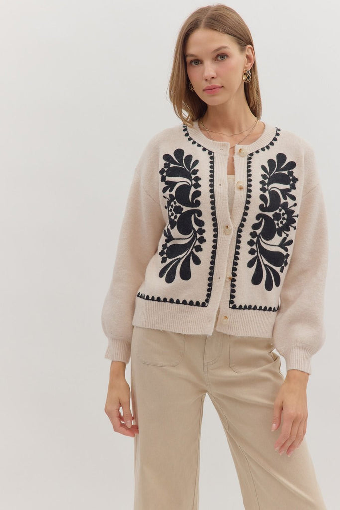 An oatmeal-colored sweater knit cardigan with black floral embroidery on the front panels and a scalloped trim on the crew neckline, hem, and button placket.