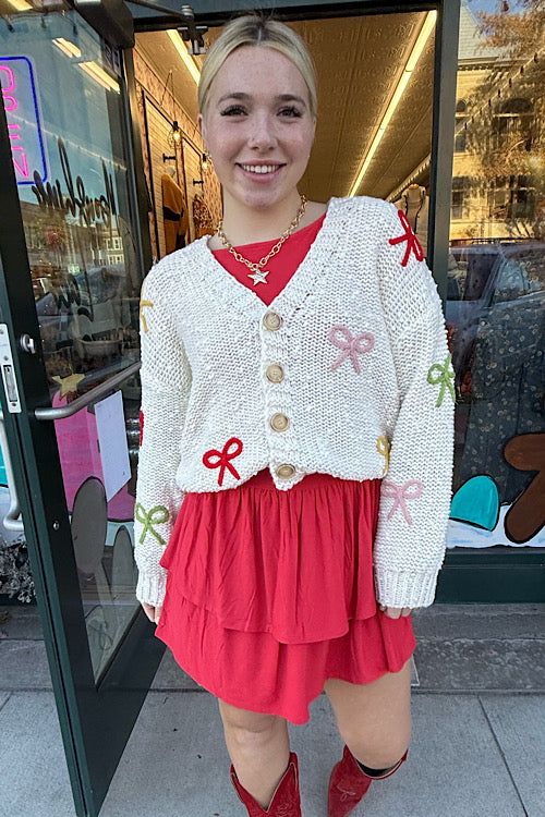 Woman wearing a white cardigan with red bows and a red skirt standing in front of a store entrance.