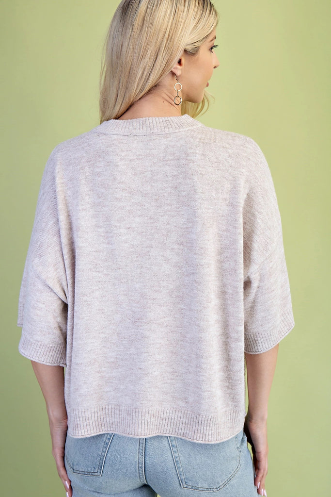 A product shot of a smiling blonde woman wearing a beige sweater with a black horse graphic on the front. She is also wearing light blue jeans and a tan wide-brim hat. The background is a solid light green.