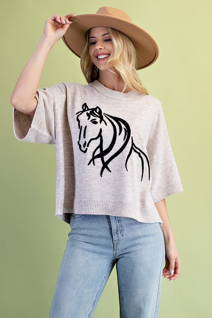 A product shot of a smiling blonde woman wearing a beige sweater with a black horse graphic on the front. She is also wearing light blue jeans and a tan wide-brim hat. The background is a solid light green.