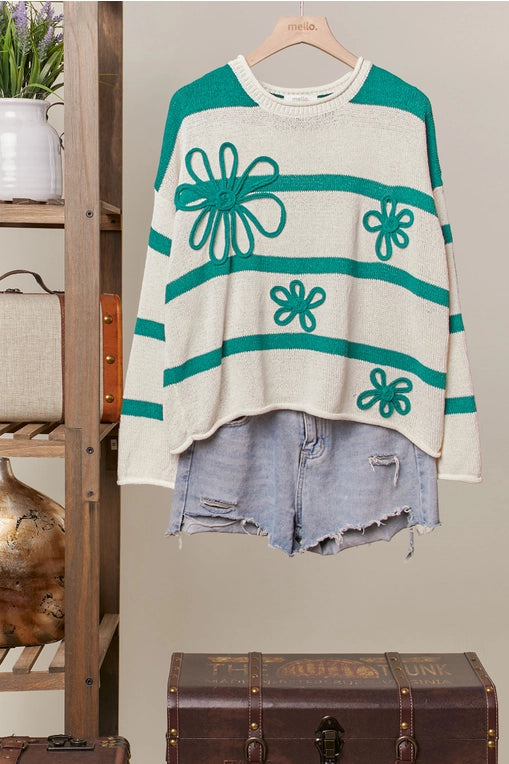 A green-and-cream striped knit sweater adorned with three-dimensional daisy flower appliques is displayed on a hanger, styled over distressed light blue denim shorts.