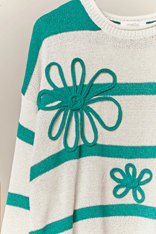 A green-and-cream striped knit sweater adorned with three-dimensional daisy flower appliques is displayed on a hanger, styled over distressed light blue denim shorts.