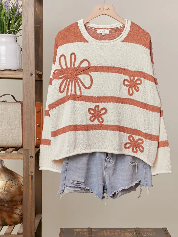 A rust-and-cream striped knit sweater with large daisy appliques is shown hanging on a clothing rack, styled with distressed denim shorts.