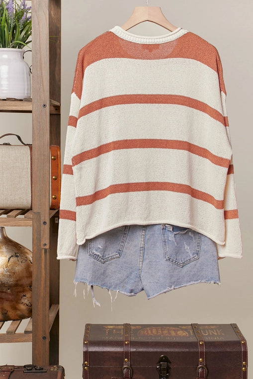 A rust-and-cream striped knit sweater with large daisy appliques is shown hanging on a clothing rack, styled with distressed denim shorts.