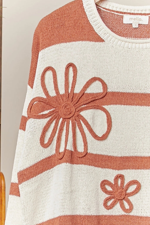 A rust-and-cream striped knit sweater with large daisy appliques is shown hanging on a clothing rack, styled with distressed denim shorts.