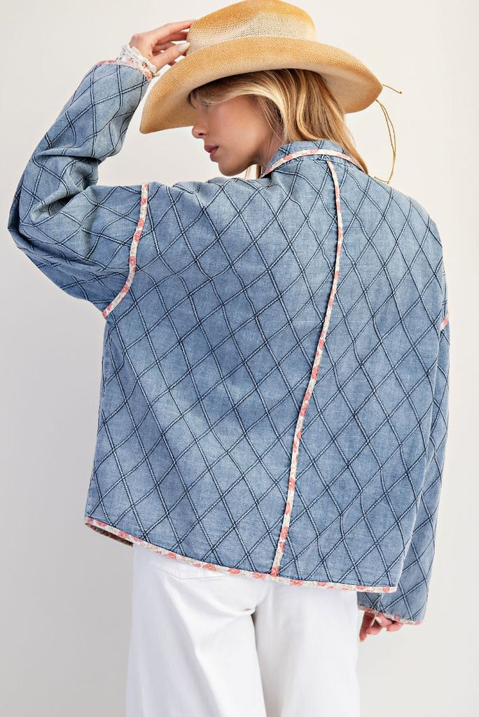 A washed denim jacket with a quilted diamond pattern. It has a pink floral print trim, large front pockets, and unique toggle or frog button closures.