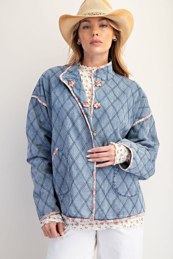 A washed denim jacket with a quilted diamond pattern. It has a pink floral print trim, large front pockets, and unique toggle or frog button closures.