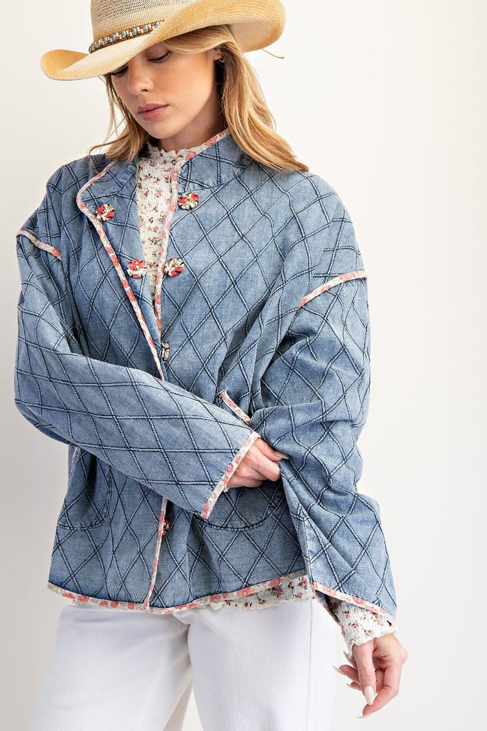 A washed denim jacket with a quilted diamond pattern. It has a pink floral print trim, large front pockets, and unique toggle or frog button closures.