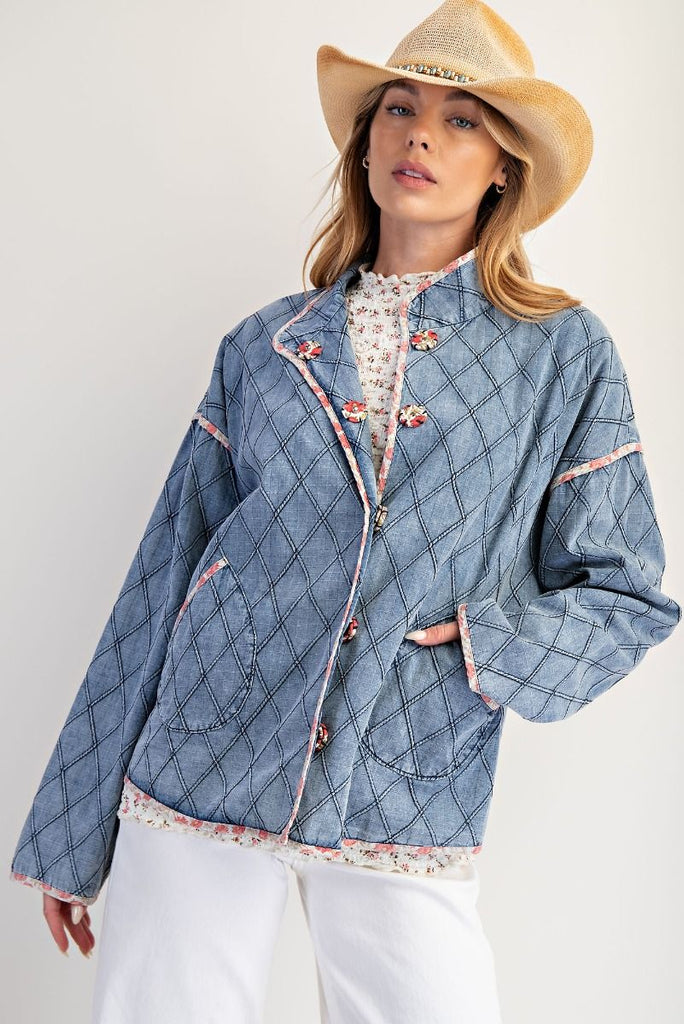 A washed denim jacket with a quilted diamond pattern. It has a pink floral print trim, large front pockets, and unique toggle or frog button closures.