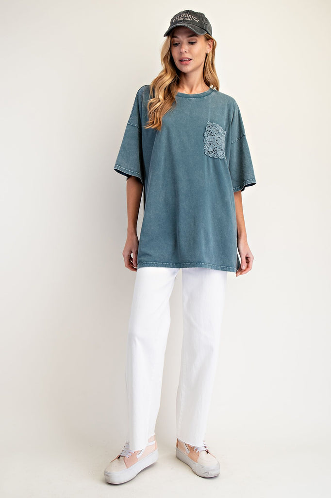 Woman wearing an oversized faded teal mineral-washed top with wide short sleeves, a crew neckline, and a square crochet pocket on the chest. Styled with white pants and a black baseball cap, the relaxed boxy silhouette gives a casual and effortless look