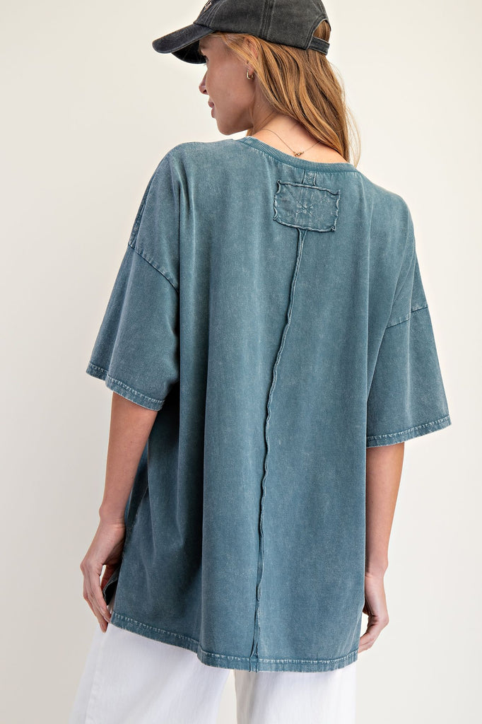 Woman wearing an oversized faded teal mineral-washed top with wide short sleeves, a crew neckline, and a square crochet pocket on the chest. Styled with white pants and a black baseball cap, the relaxed boxy silhouette gives a casual and effortless look