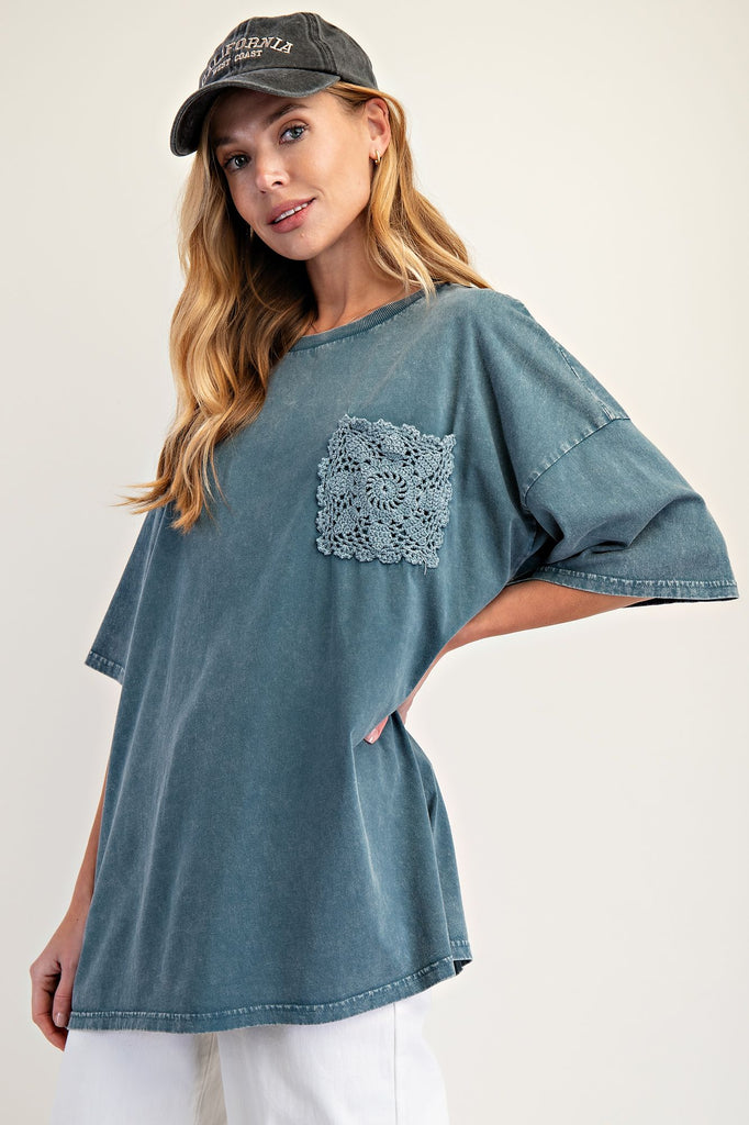 Woman wearing an oversized faded teal mineral-washed top with wide short sleeves, a crew neckline, and a square crochet pocket on the chest. Styled with white pants and a black baseball cap, the relaxed boxy silhouette gives a casual and effortless look