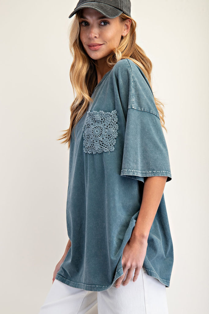 Woman wearing an oversized faded teal mineral-washed top with wide short sleeves, a crew neckline, and a square crochet pocket on the chest. Styled with white pants and a black baseball cap, the relaxed boxy silhouette gives a casual and effortless look