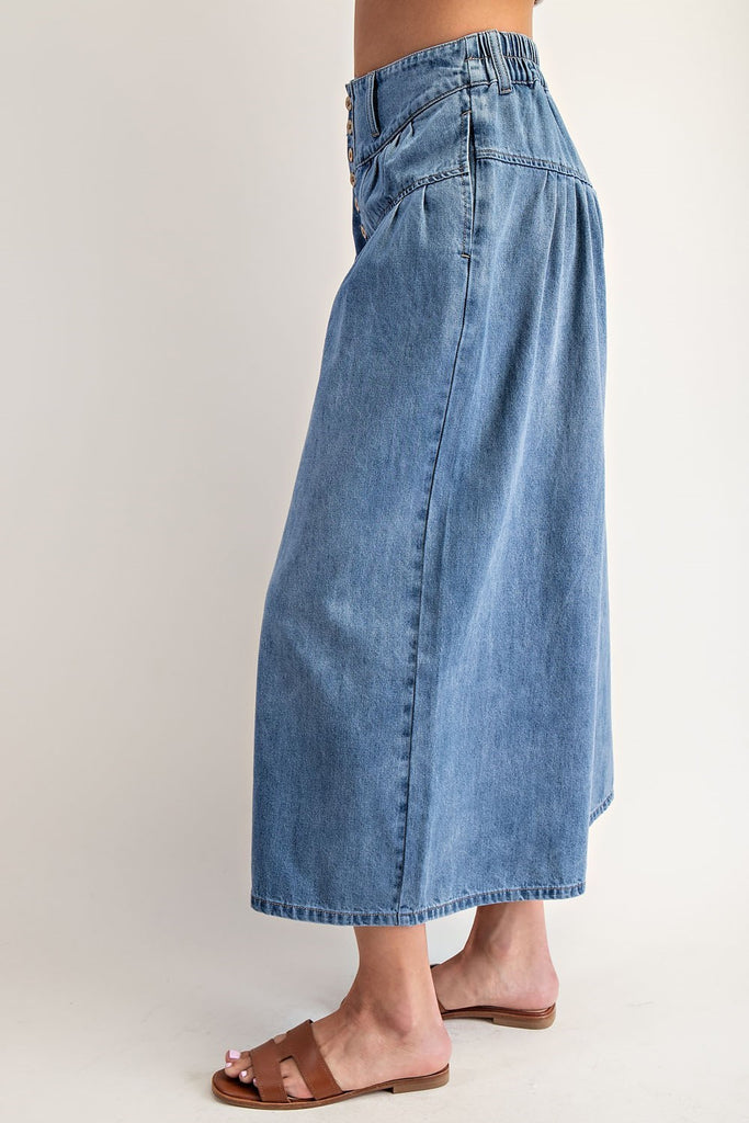 Close-up of a woman wearing high-rise stone-washed denim culottes with a button fly, pleated waist, wide cropped legs, and brown leather slide sandals.