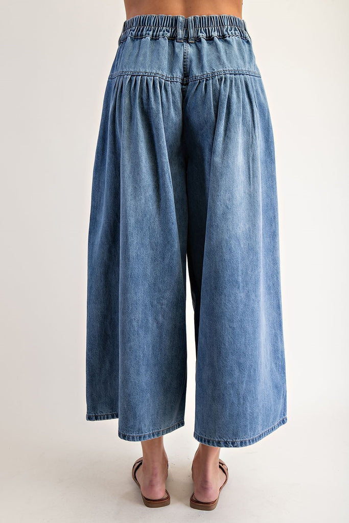Close-up of a woman wearing high-rise stone-washed denim culottes with a button fly, pleated waist, wide cropped legs, and brown leather slide sandals.