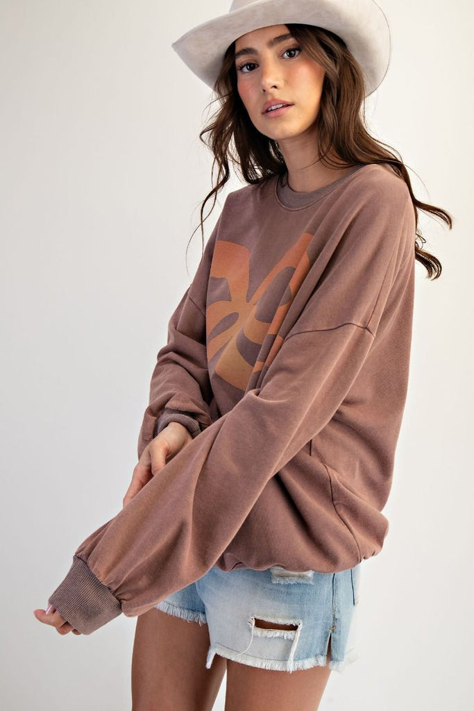 A mocha-colored, oversized pullover made of washed terry fabric. It has a large orange and tan bow graphic on the front, a crew neckline, and long sleeves.