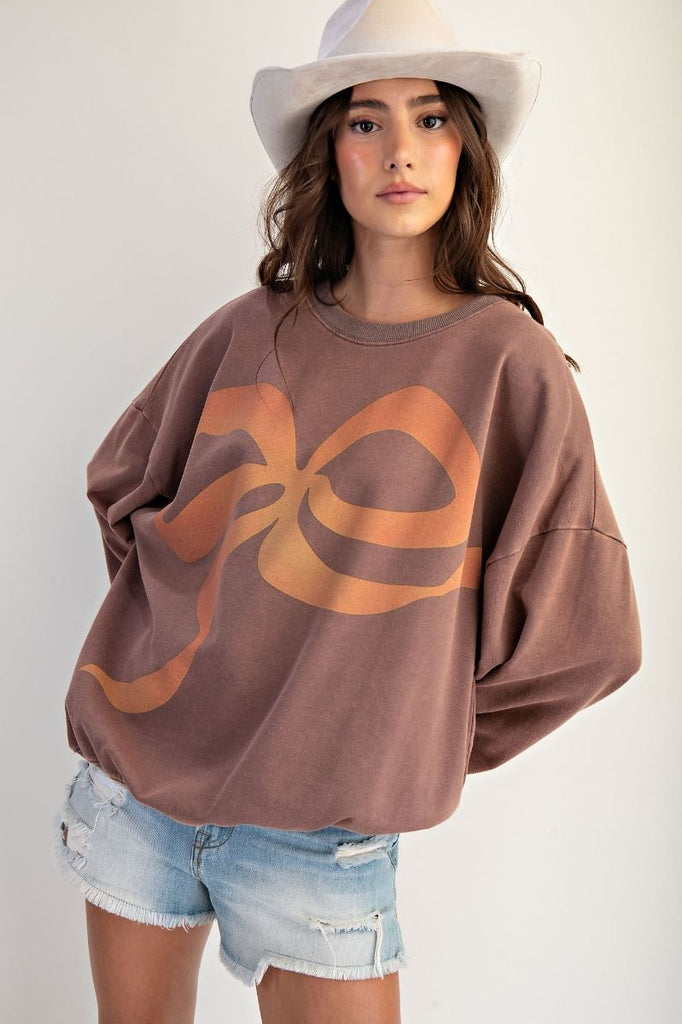 A mocha-colored, oversized pullover made of washed terry fabric. It has a large orange and tan bow graphic on the front, a crew neckline, and long sleeves.