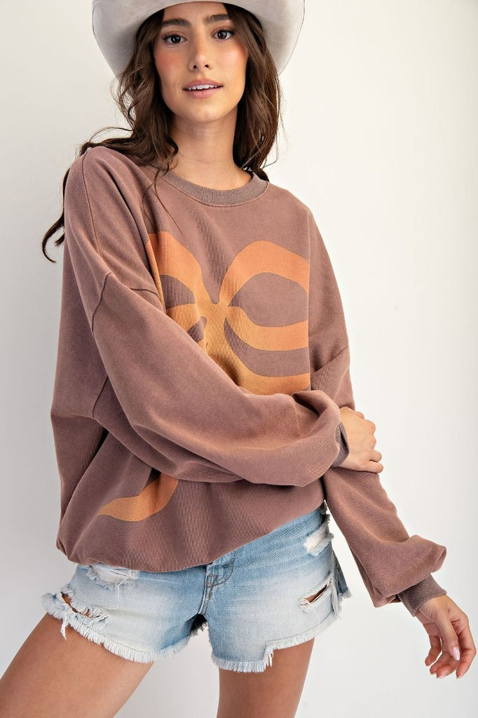 A mocha-colored, oversized pullover made of washed terry fabric. It has a large orange and tan bow graphic on the front, a crew neckline, and long sleeves.