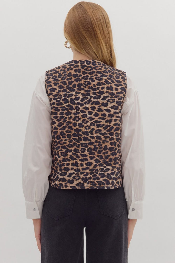A woman is wearing a sleeveless, quilted leopard print vest with a self-tie front that creates a large bow. She is layering the vest over a white long-sleeve shirt.