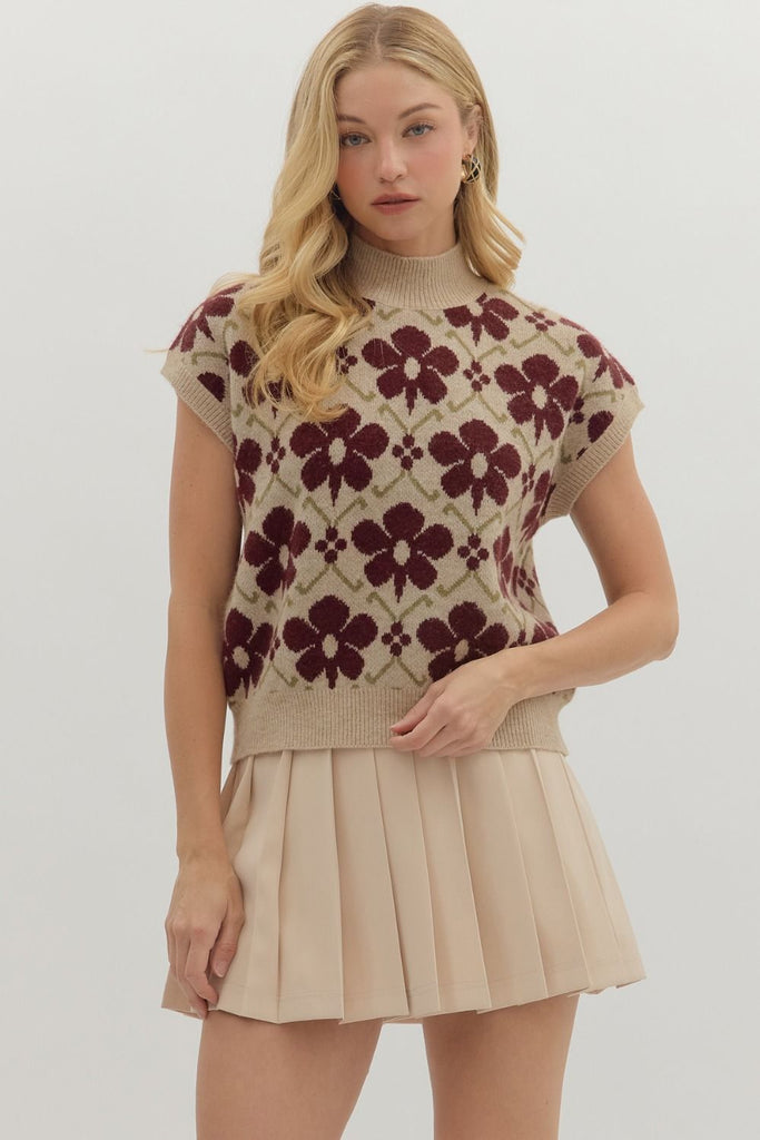 A woman wearing a sleeveless beige knit top with a burgundy floral pattern and high neckline, paired with a pleated beige mini skirt.