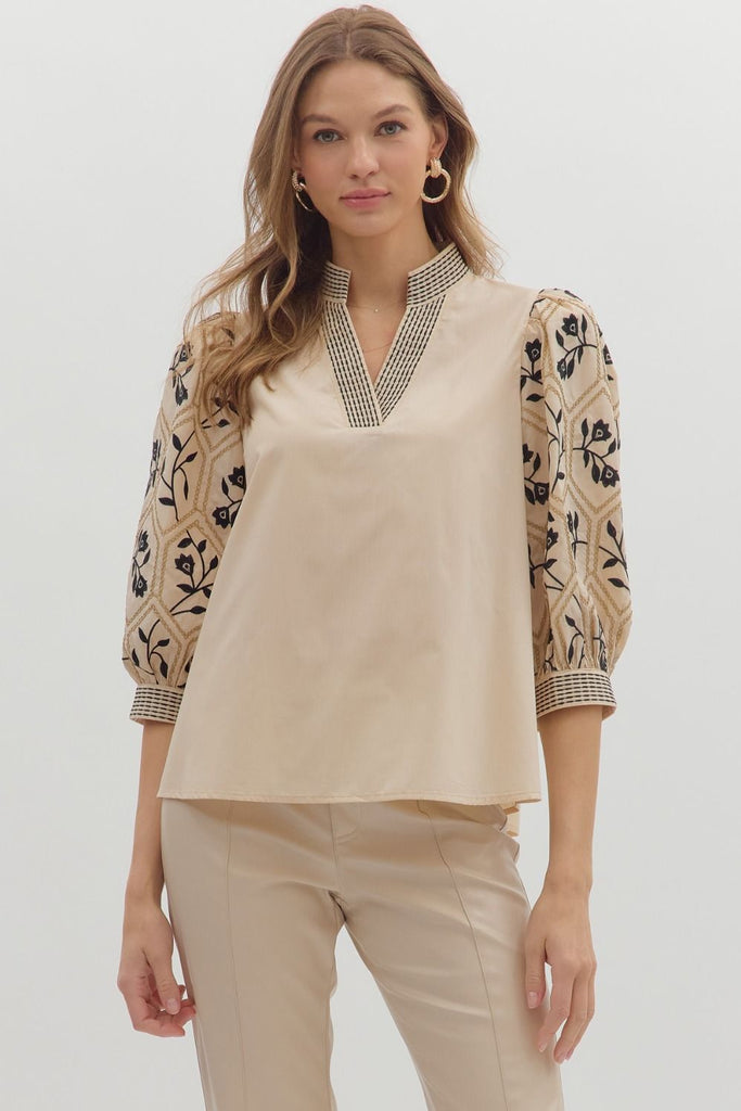 A woman wears a beige woven top with a solid-colored body and patterned, embroidered three-quarter length sleeves, featuring a geometric and floral design. The V-neck has an embroidered band.