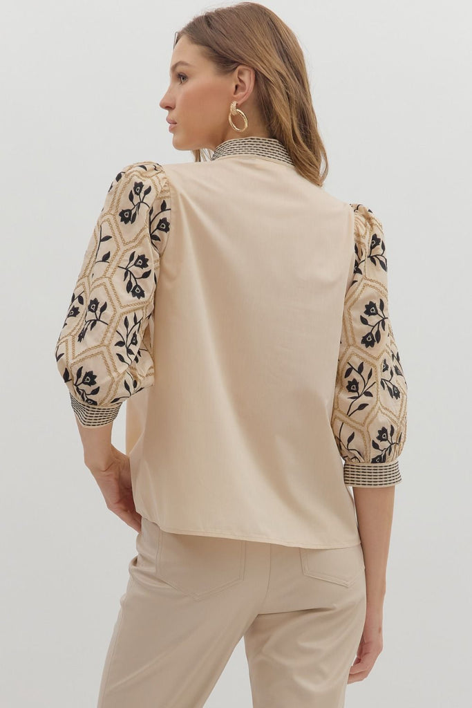  A woman wears a beige woven top with a solid-colored body and patterned, embroidered three-quarter length sleeves, featuring a geometric and floral design. The V-neck has an embroidered band.