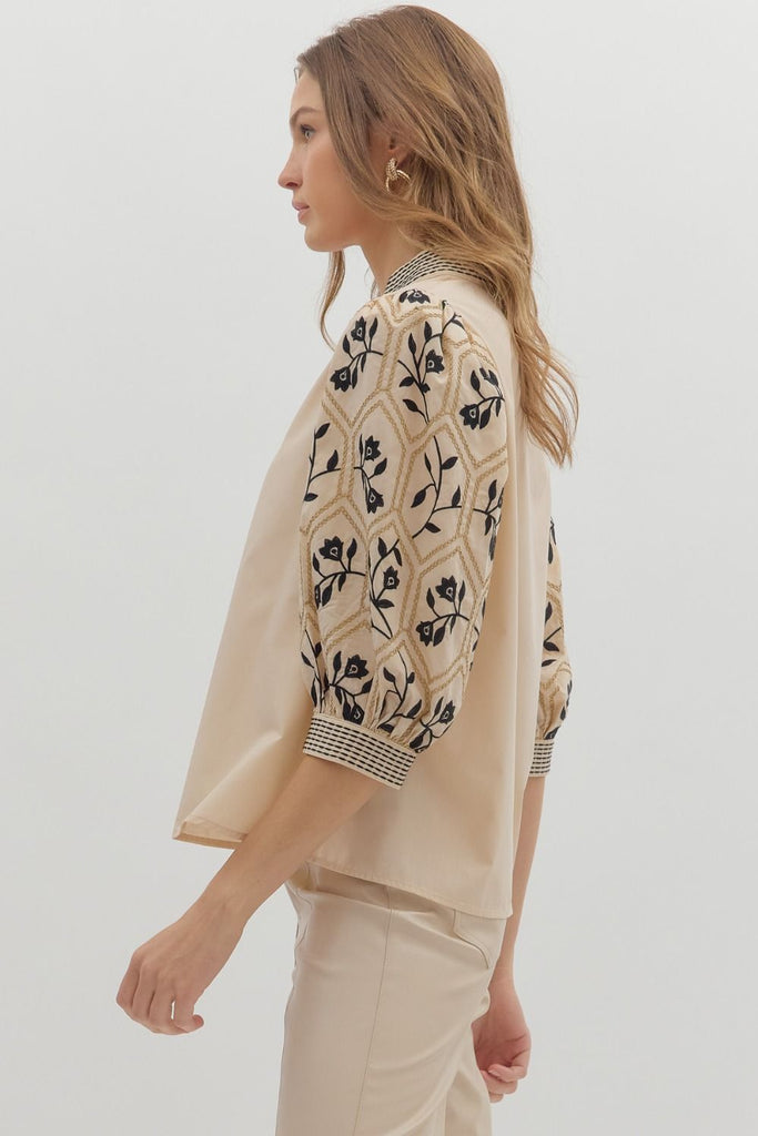 A woman wears a beige woven top with a solid-colored body and patterned, embroidered three-quarter length sleeves, featuring a geometric and floral design. The V-neck has an embroidered band.