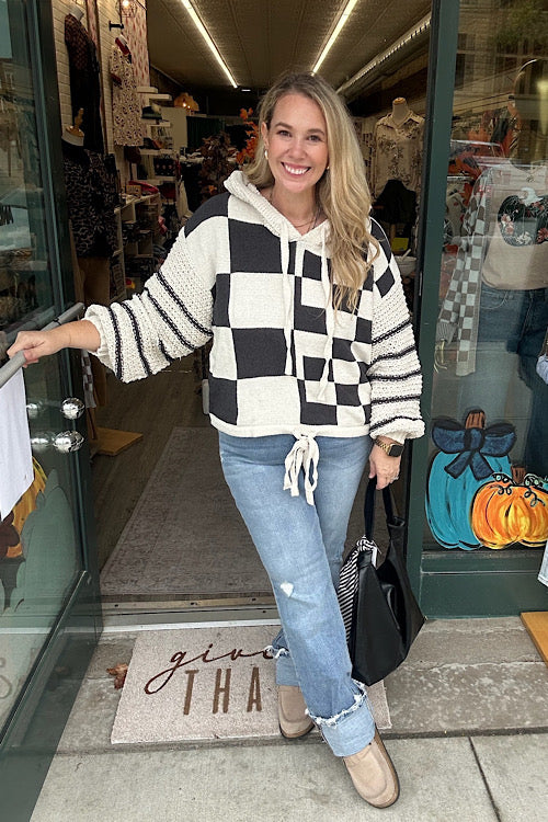 A woman models a hooded, black and off-white checkerboard sweater with textured, contrasting knit sleeves and a drawstring hem, styled with denim jeans in a store setting.