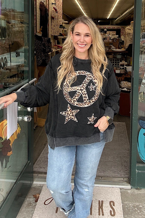 Woman models Euphemia leopard peace sign sweatshirt in washed black with a large animal print graphic, a relaxed fit, and distressed raw hem with denim jeans in store setting. 