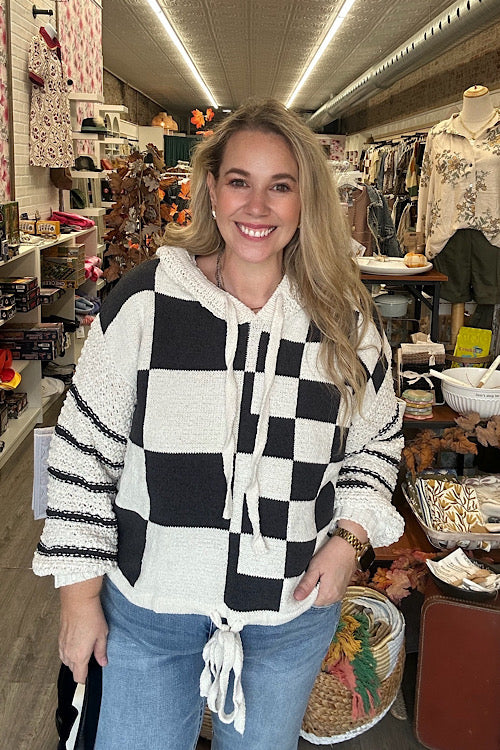 A woman models a hooded, black and off-white checkerboard sweater with textured, contrasting knit sleeves and a drawstring hem, styled with denim jeans in a store setting.
