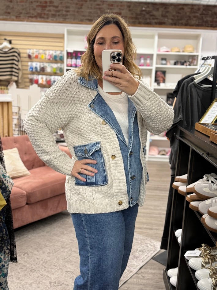 Woman taking a mirror selfie wearing a white cardigan with denim accents in a store.