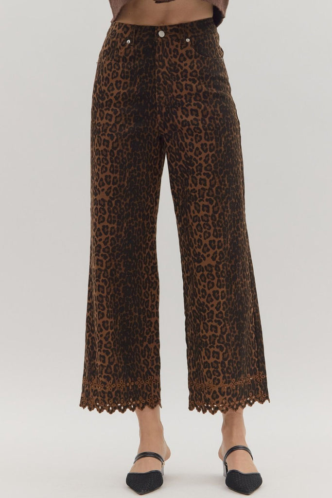 A close-up shot of the lower legs of a person wearing leopard print, wide-leg cropped pants with a floral embroidered and scalloped hemline. They are styled with black mules.