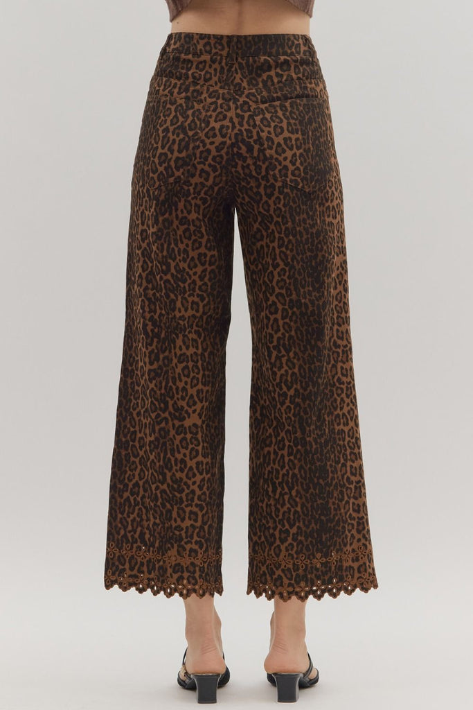 A close-up shot of the lower legs of a person wearing leopard print, wide-leg cropped pants with a floral embroidered and scalloped hemline. They are styled with black mules.