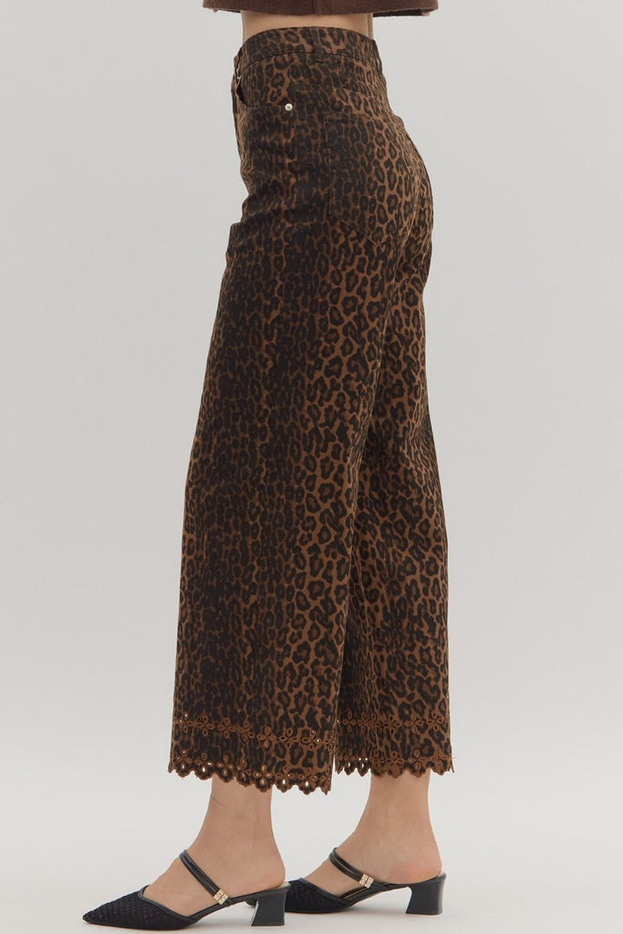 A close-up shot of the lower legs of a person wearing leopard print, wide-leg cropped pants with a floral embroidered and scalloped hemline. They are styled with black mules.
