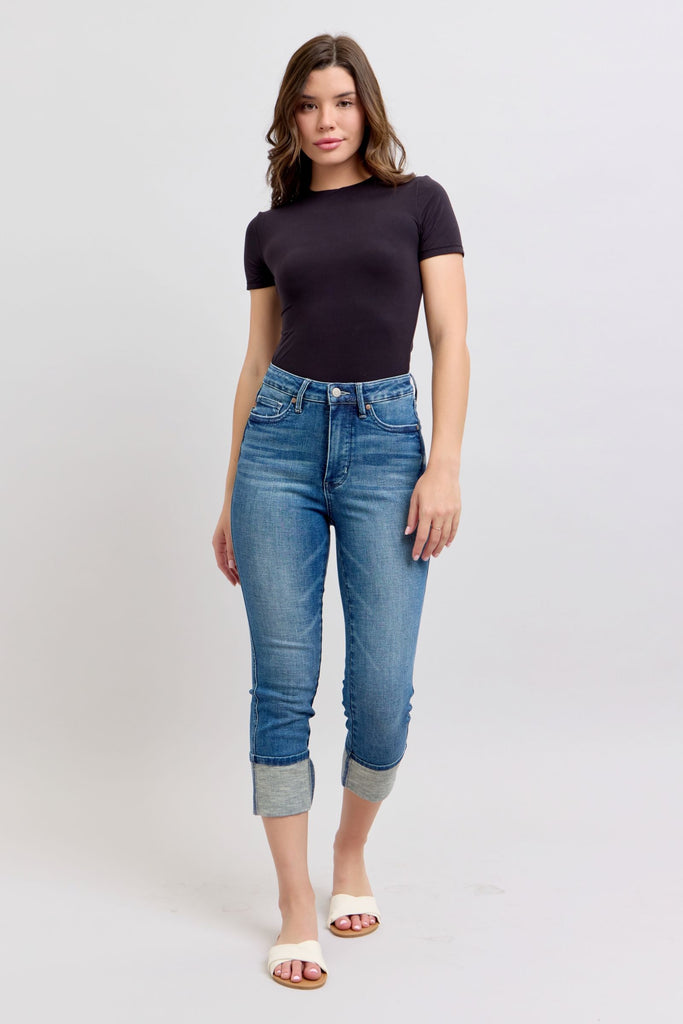 Eva - Judy Blue Tummy Control Cuff Capri Jeans shown styled with black fitted t-shirt. Medium wash denim features high waist design, cuffed capri length, and classic 5-pocket styling. Model wears jeans with white sandals against light background.