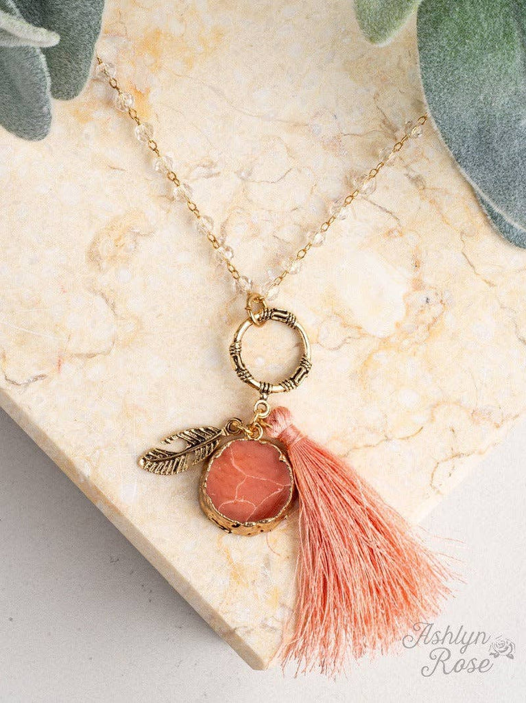 Peach beaded necklace with gold feather charm, round peach stone, and tassel accent.