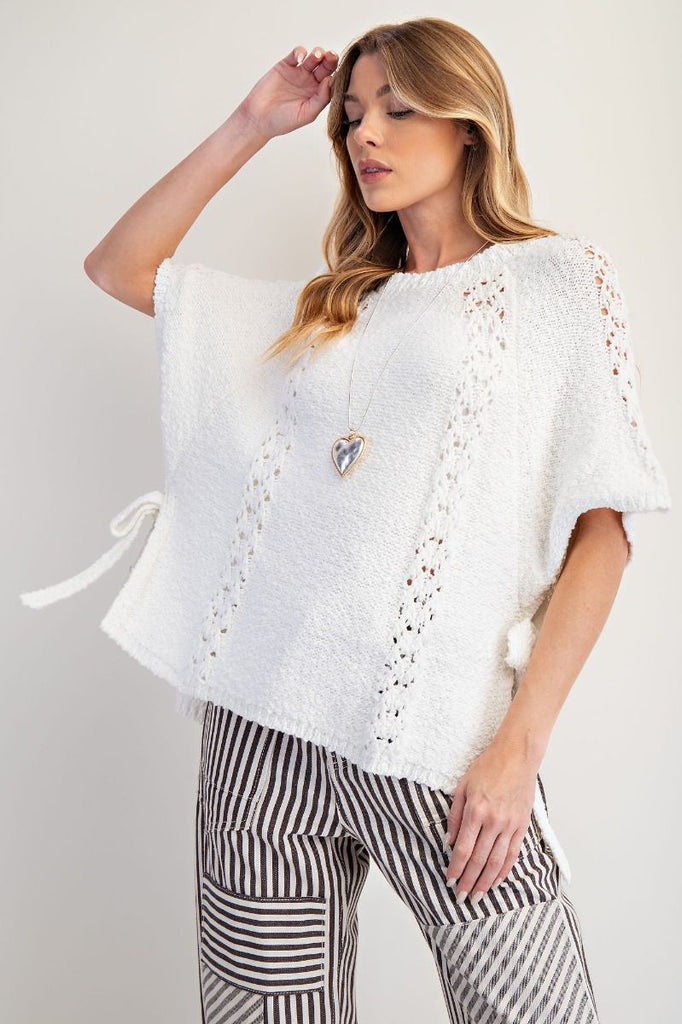 Off-white knitted poncho sweater with short sleeves featuring crochet-style lace panels, crew neckline, relaxed boxy fit, and open sides with adjustable ties