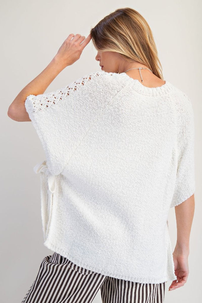 Off-white knitted poncho sweater with short sleeves featuring crochet-style lace panels, crew neckline, relaxed boxy fit, and open sides with adjustable ties