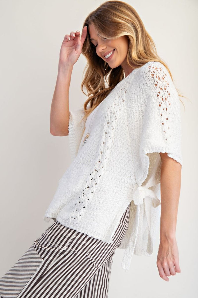 Off-white knitted poncho sweater with short sleeves featuring crochet-style lace panels, crew neckline, relaxed boxy fit, and open sides with adjustable ties