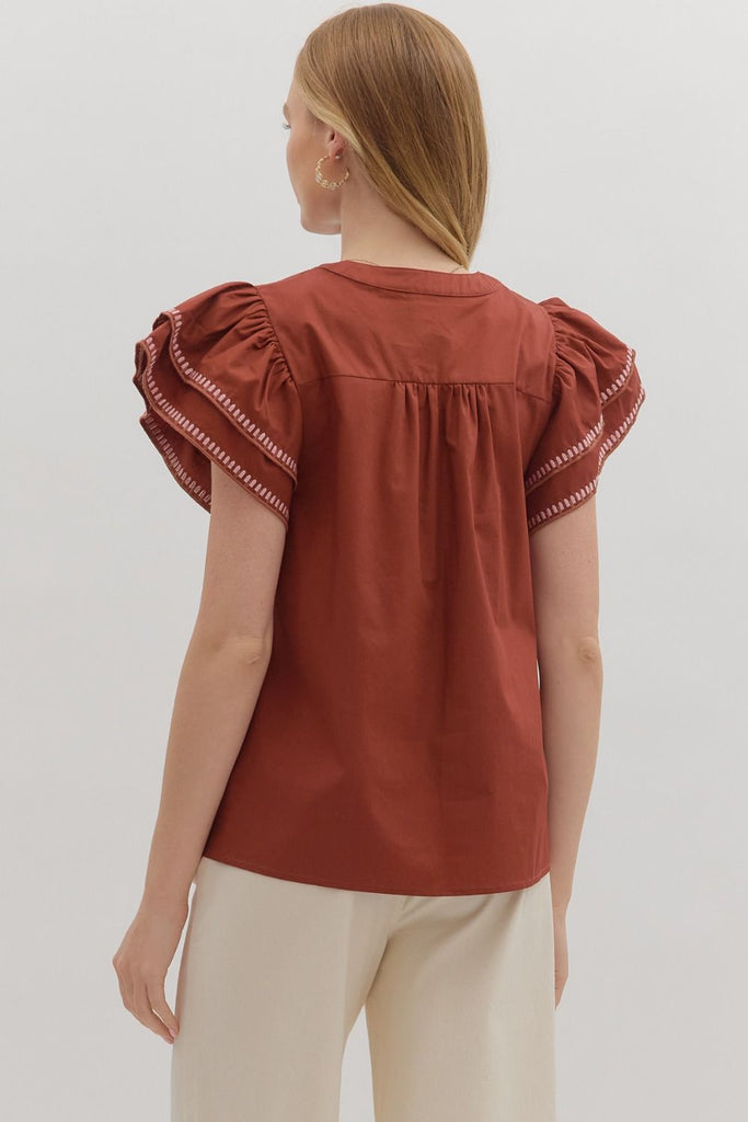 A solid brick-red top with tiered ruffle flutter sleeves. The v-neckline and sleeve ruffles have a white contrast embroidered stitching.