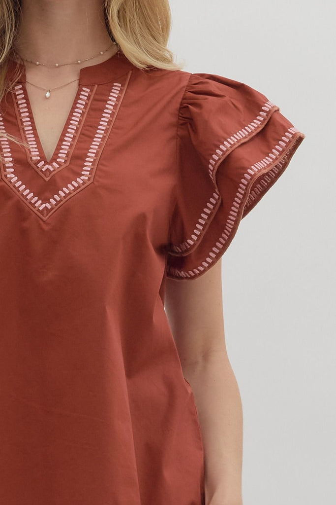 A solid brick-red top with tiered ruffle flutter sleeves. The v-neckline and sleeve ruffles have a white contrast embroidered stitching.