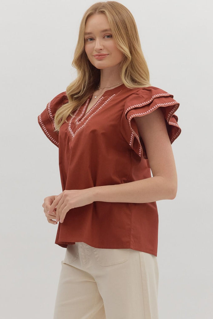 A solid brick-red top with tiered ruffle flutter sleeves. The v-neckline and sleeve ruffles have a white contrast embroidered stitching.