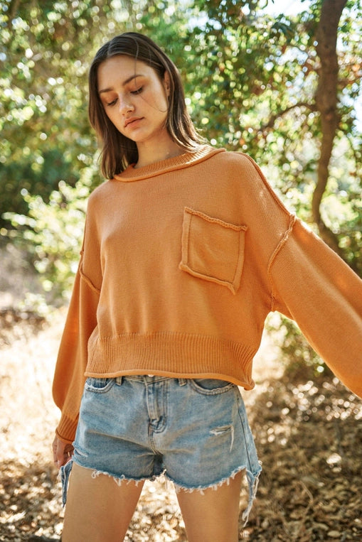 Woman wearing a loose-fitting tan round neck sweater with dropped shoulders, a front chest pocket, and ribbed hem and cuffs, paired with distressed light-wash denim shorts, standing outdoors among sunlit trees.