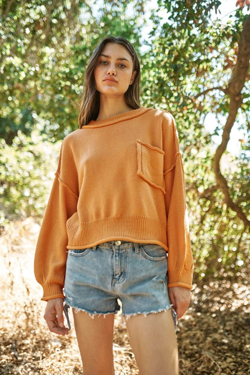 Woman wearing a loose-fitting tan round neck sweater with dropped shoulders, a front chest pocket, and ribbed hem and cuffs, paired with distressed light-wash denim shorts, standing outdoors among sunlit trees.