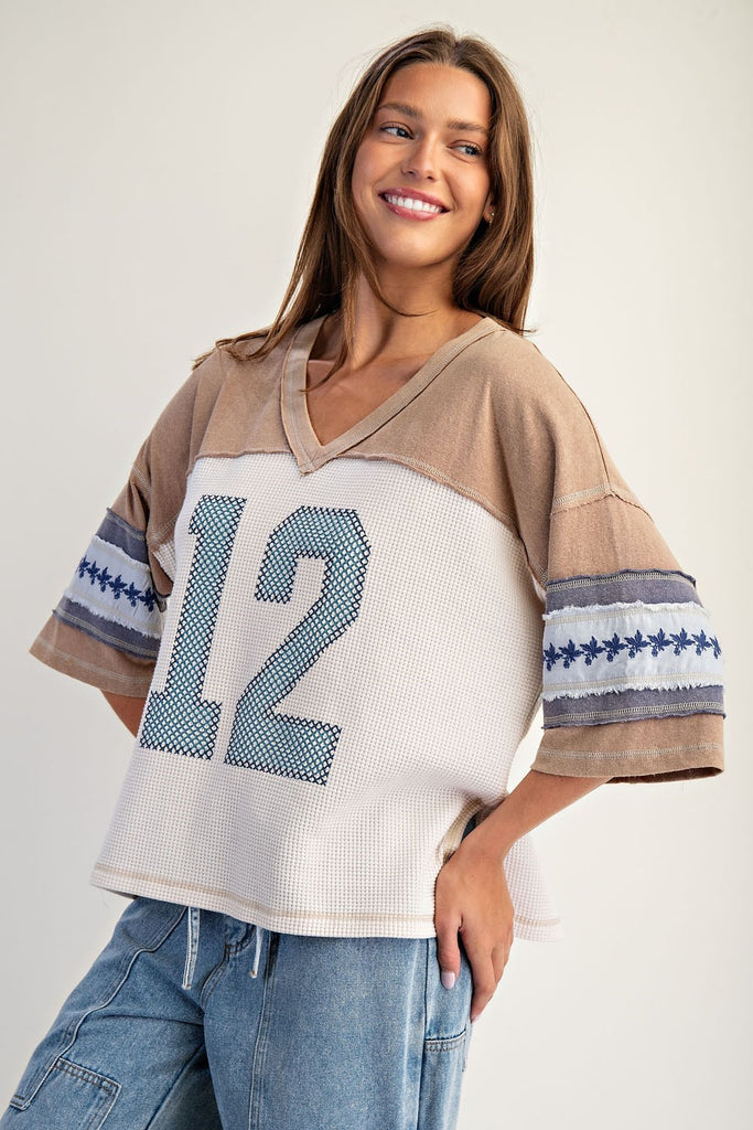 Model wearing a snow-washed waffle knit varsity t-shirt with beige jersey sleeves, large number 12 graphic in blue at front, and embroidered floral detailing on the wide short sleeves. Styled with relaxed denim bottoms.
